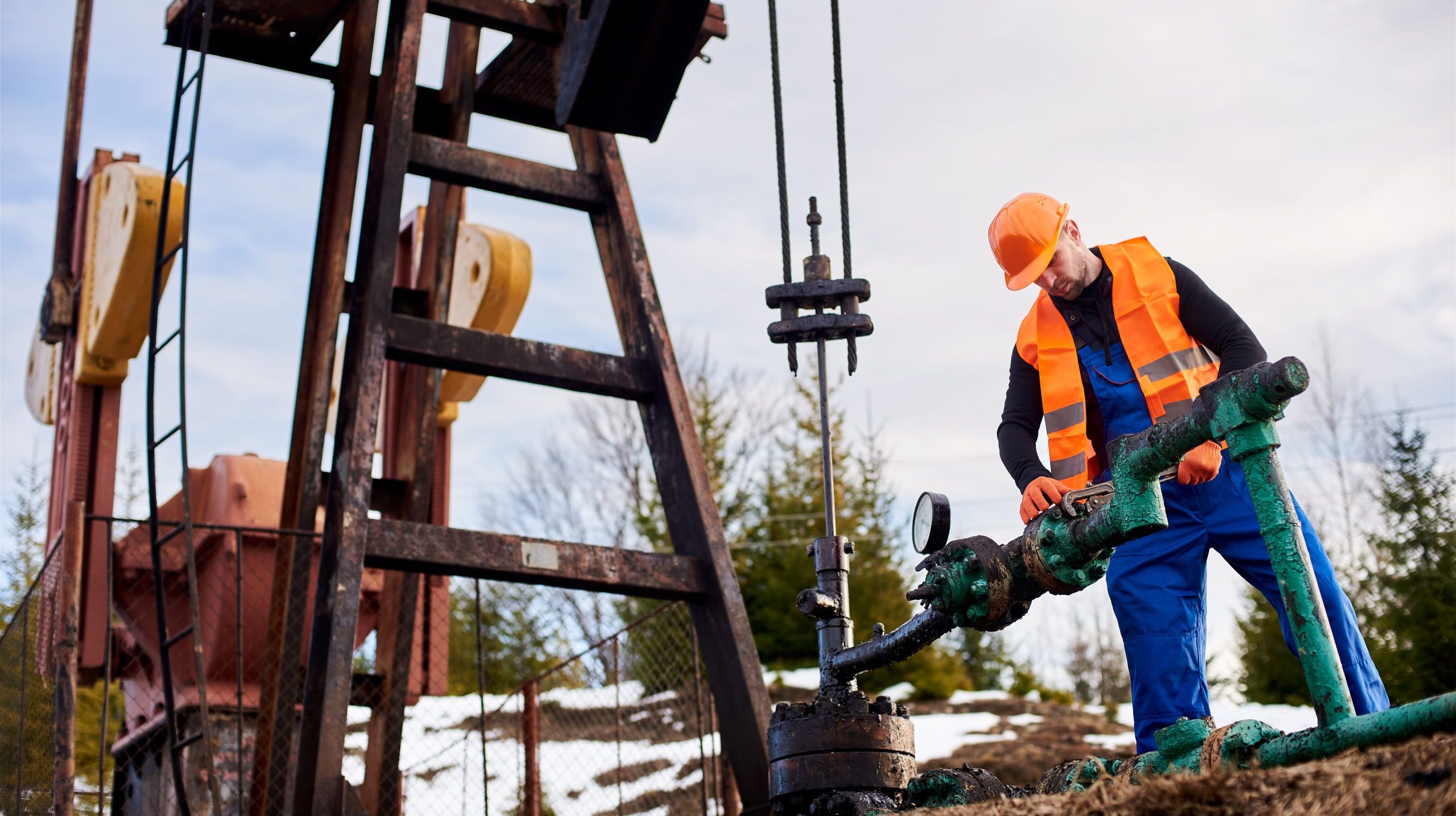 Oil worker in orange uniform and helmet working with a pipe wrench near an oil pump jack Oil worker in blue overalls, orange vest and helmet using industrial wrench while repairing pipe of oil well pump jack, tightening bolt on pipe. Concept of petroleum industry and repair.
